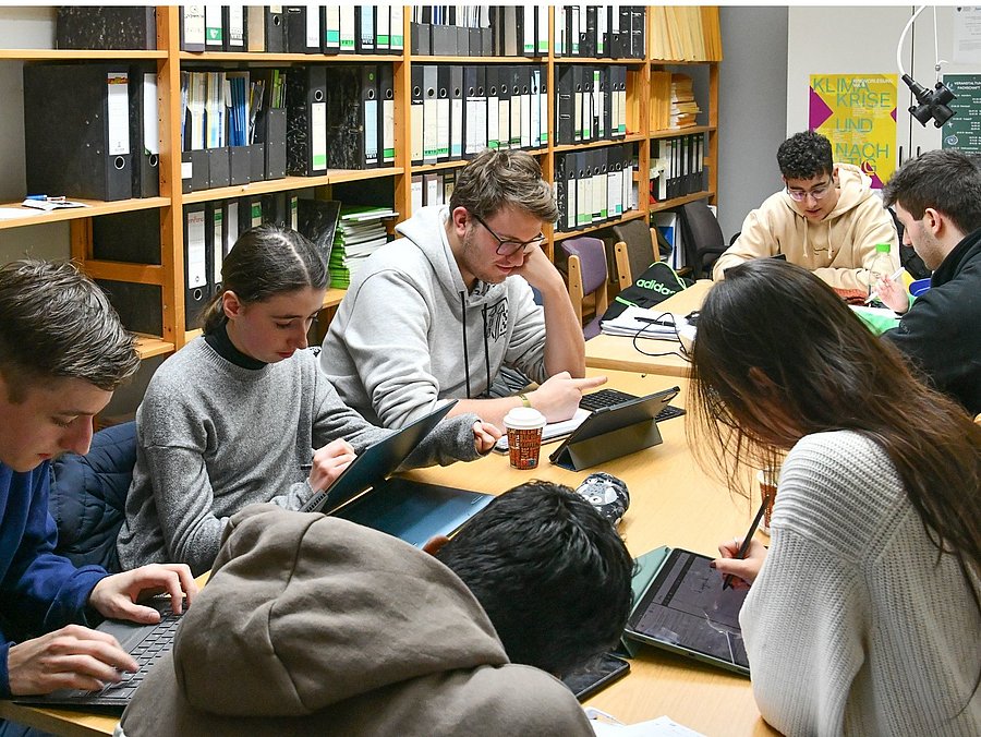 Gezeigt wird ein Foto mit mehreren Studierenden der Physik, welche an einem Gruppentisch sitzend arbeiten.