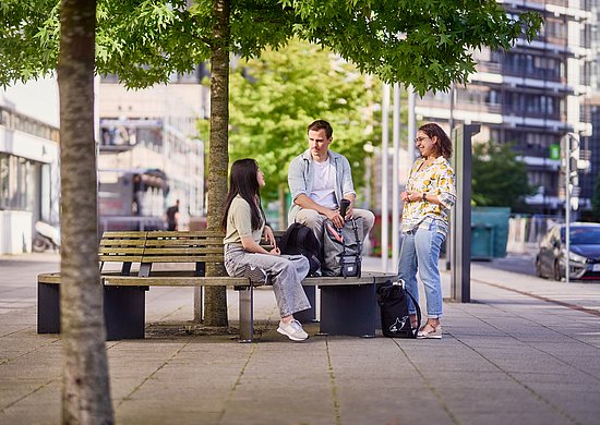 Gezeigt wird ein Image-Foto von 3 Studierenden, welche sich vor dem Gebäude VW miteinander unterhalten.